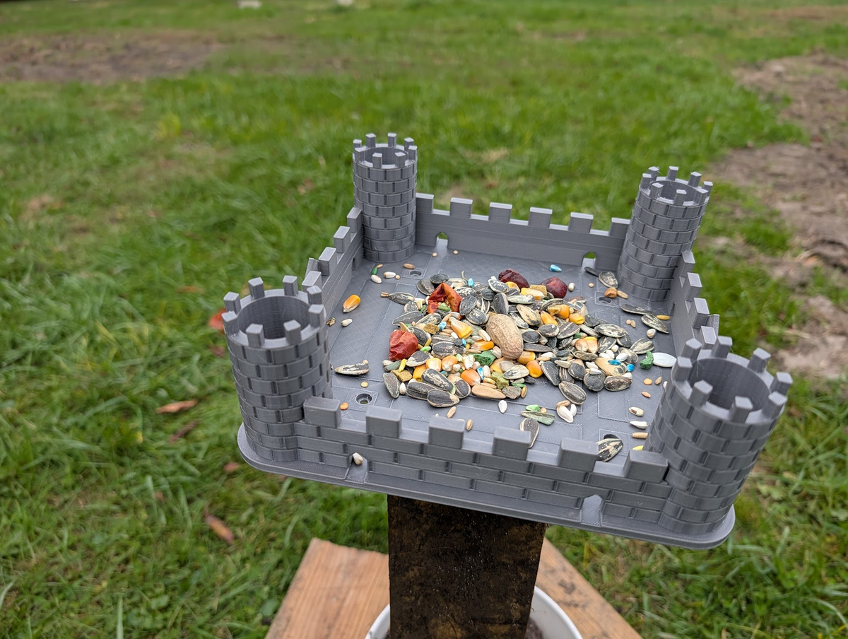Grey medieval castle bird feeder with seed in the open tray, mounted on a wooden post